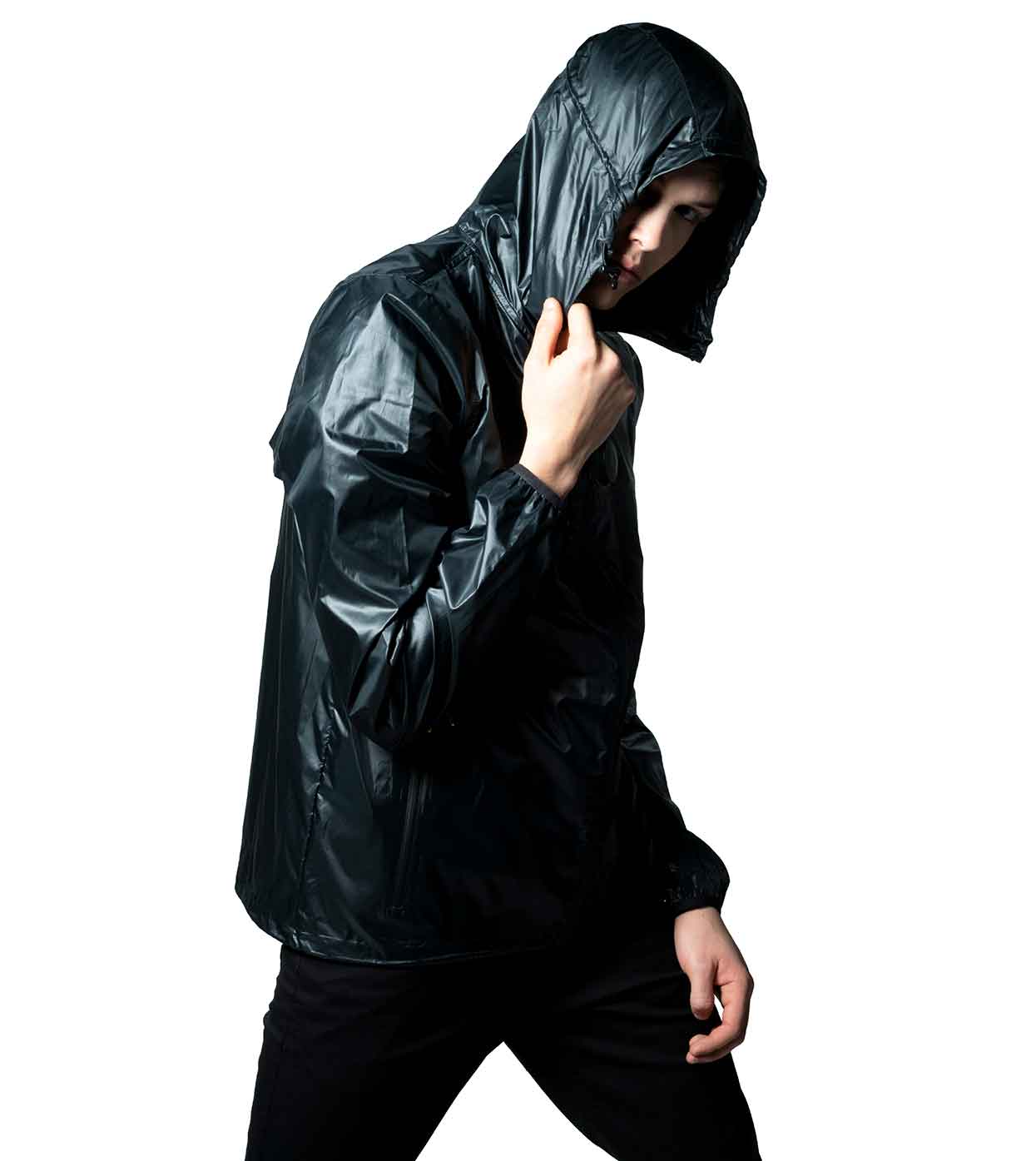 Clad in Machina - Wearable Technology's BLACK WINDBREAKER, a glossy black hooded jacket made from ultralight nylon, a person draws the hood over their head. Their intense look is accentuated against the simple white background, creating an aura of mystery. This blend of stylish design and fashion innovation results in a captivating image.