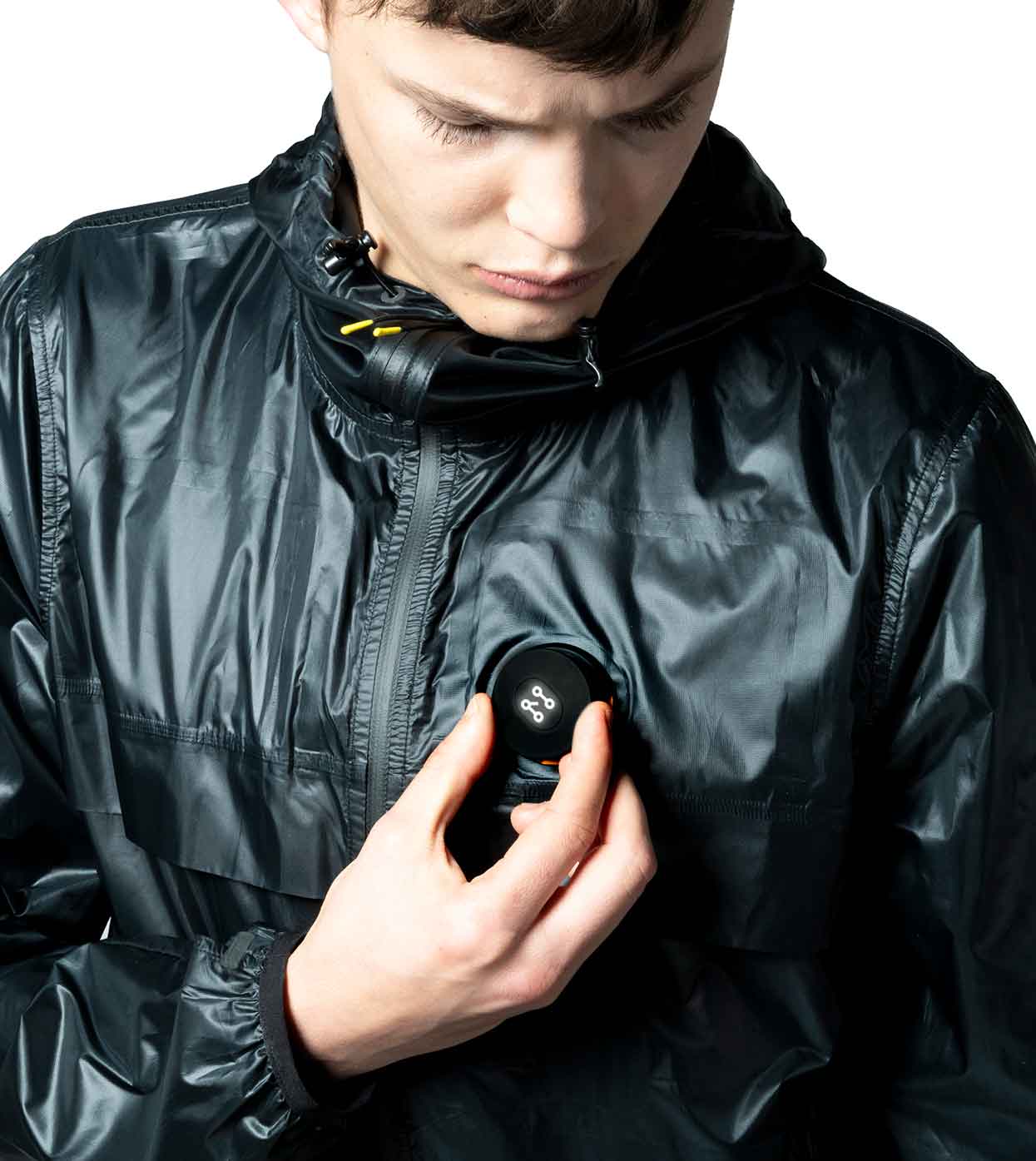 A person is wearing a BLACK WINDBREAKER by Machina - Wearable Technology, made of shiny, ultralight nylon. They are looking down at a round gadget fastened to the jacket, which features buttons and a display screen that highlight its fashion-forward technology against a plain white background.