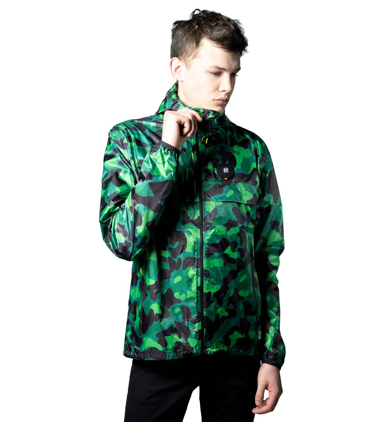 A young man models the GREEN-CAMO WINDBREAKER from Machina - Wearable Technology against a white backdrop. Holding the collar with his right hand, he gazes sideways to emphasize the jacket's innovative fashion technology. Paired with black pants, his expression is one of contemplation.