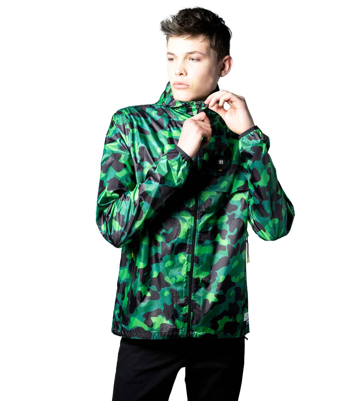 A person wearing the GREEN-CAMO WINDBREAKER by Machina - Wearable Technology is adjusting the collar. The white background contrasts with the jacket's camouflage design, crafted with fashion technology to maintain a stylish edge as the wearer looks sideways with a neutral expression.