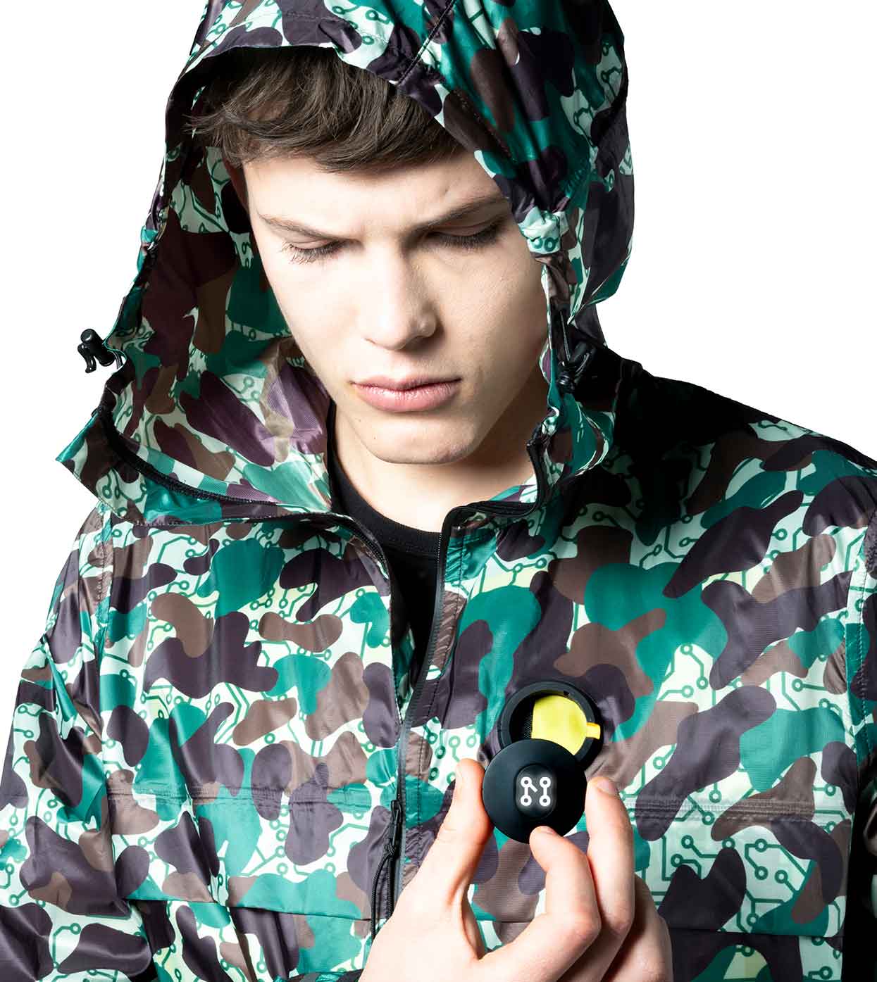 Wearing a Machina - Wearable Technology JARHEAD CAMO WINDBREAKER, an individual peers into a small round case in their hand. The case has a yellow interior and a sleek black exterior adorned with a white symbol on top. Set against a plain white background, this scene accentuates the fusion of fashion technology and style.
