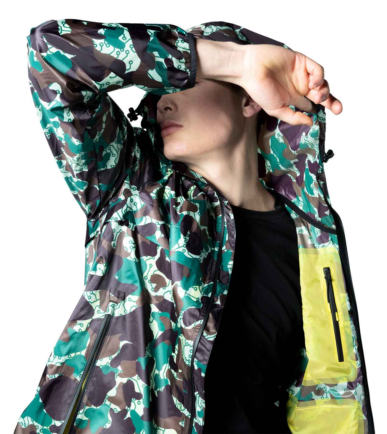 A model showcases the JARHEAD CAMO WINDBREAKER by Machina - Wearable Technology, an ultralight nylon hooded jacket featuring a green, brown, and black camouflage pattern. The smart windbreaker includes a distinctive yellow zippered pocket, perfectly merging fashion and technology against a white backdrop.