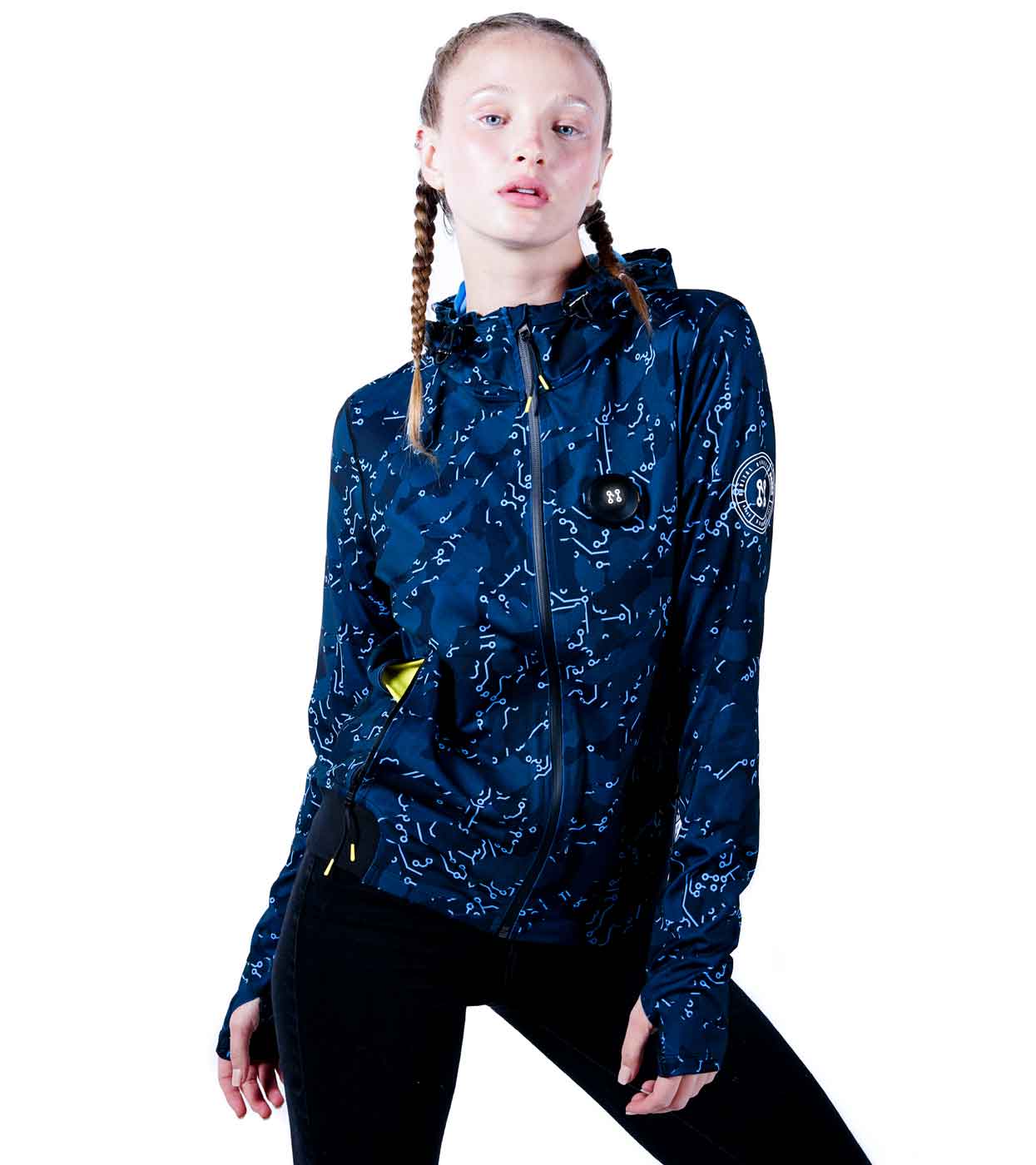 A person with braided hair wearing the ELECTRIC BLUE HOODIE from Machina - Wearable Technology stands against a white background, looking directly at the camera. The hoodie, featuring a circular logo on the sleeve and yellow details at the zippers, closely resembles an advanced jacket.