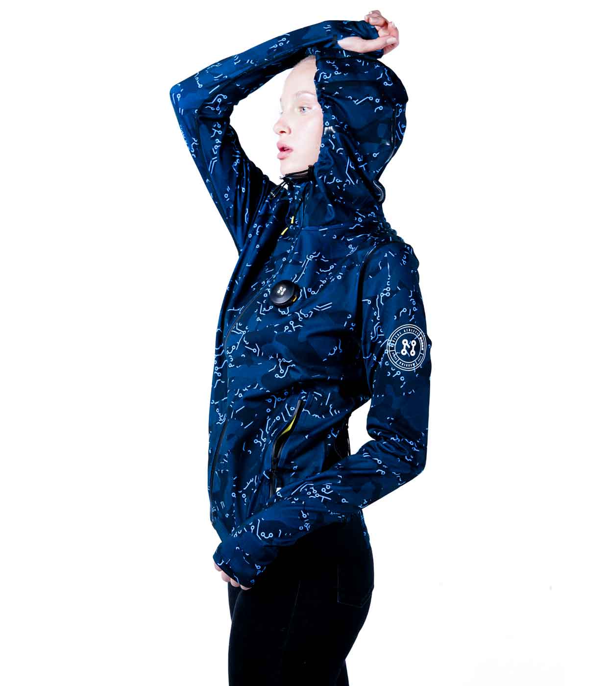 A person with a light skin tone is wearing the ELECTRIC BLUE HOODIE by Machina - Wearable Technology. They are standing in side profile with one arm raised, touching their forehead. The hoodie, featuring circular patches and a zippered front, seamlessly blends fashion tech against the white background.